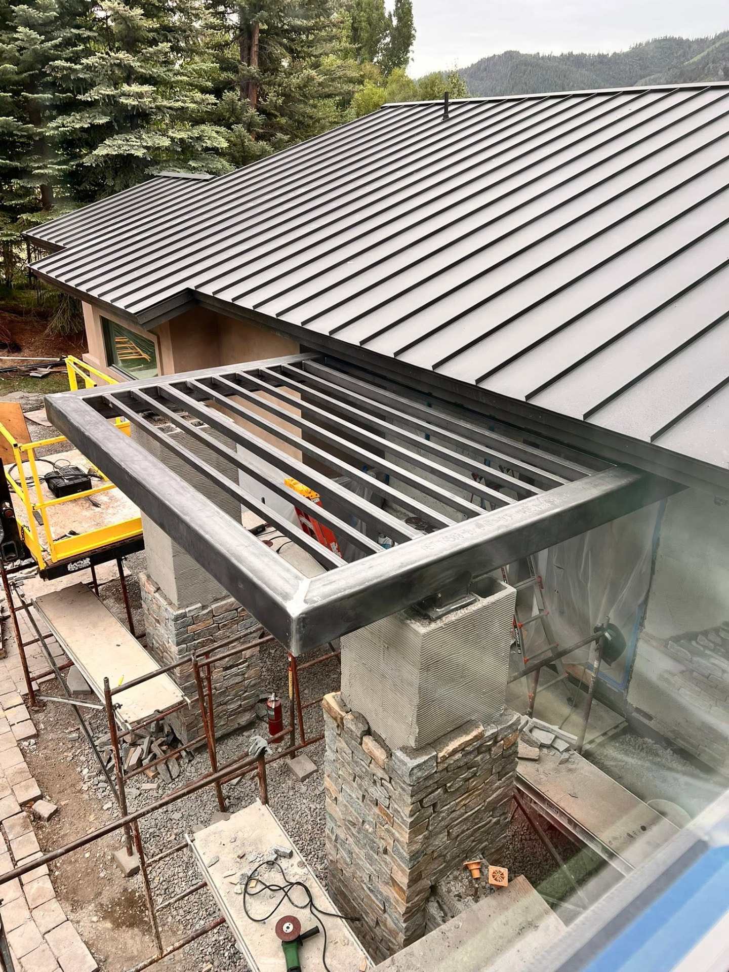 Raw welded steel pergola frame installed on stone chimney columns beside a standing-seam metal roof